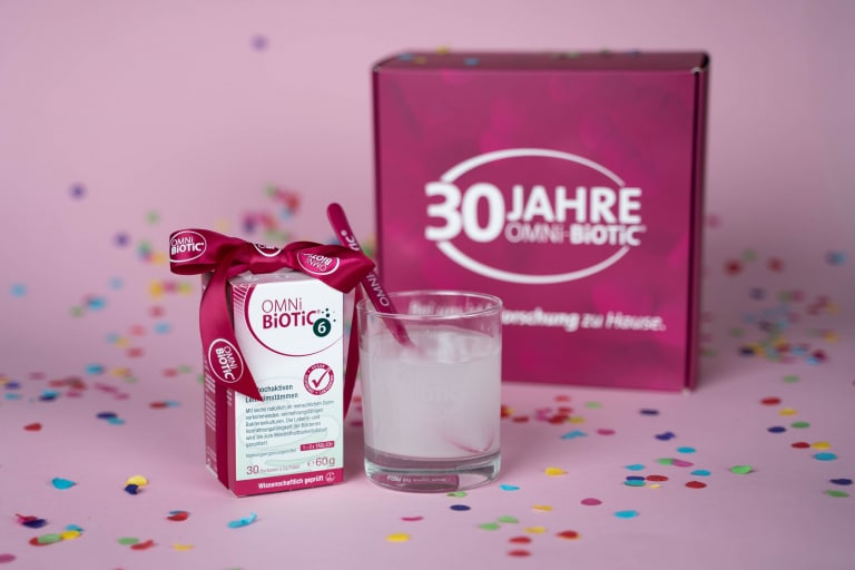 Produktfoto von OMNi-BiOTiC® 6, dekoriert mit einer Jubiläumsschleife, einem Glas mit angerührtem Produkt und einer Geschenkbox mit der Aufschrift „30 Jahre OMNi-BiOTiC®“ auf einem rosafarbenen Hintergrund mit Konfetti.